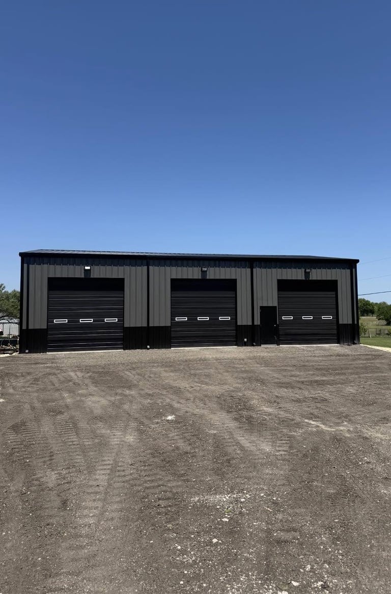 Large metal garage building with three black roll-up doors and corrugated metal siding on a gravel lot under clear blue sky