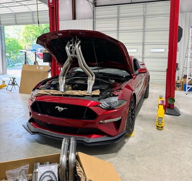 Red Mustang with open hood showing engine headers in automotive workshop with red lift