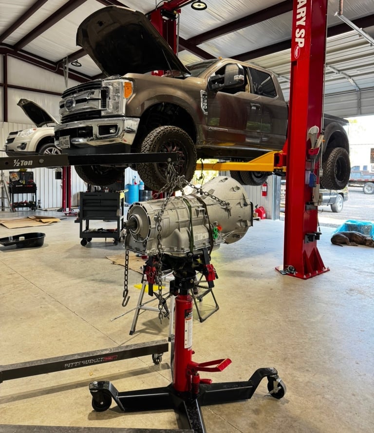 Transmission engine suspended on red hydraulic lift in auto repair shop with truck on adjacent lift