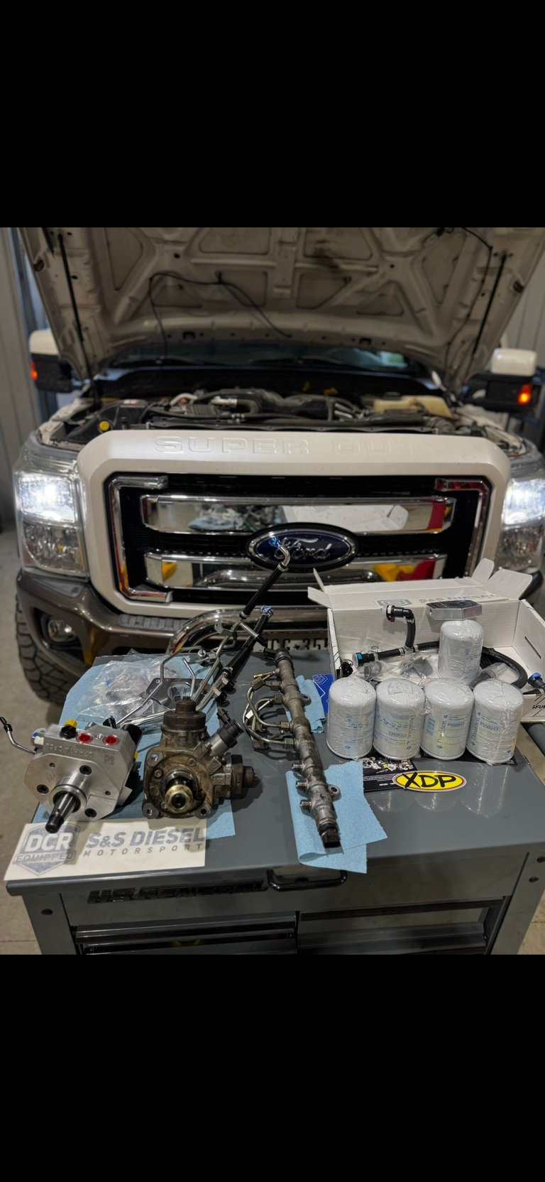 Ford diesel engine parts displayed on workbench with vehicle hood open in background showing engine bay