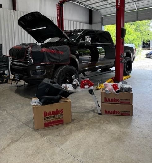 Black pickup truck in garage with hood open, engine compartment visible, Banks branded boxes and performance parts displayed on concrete floor